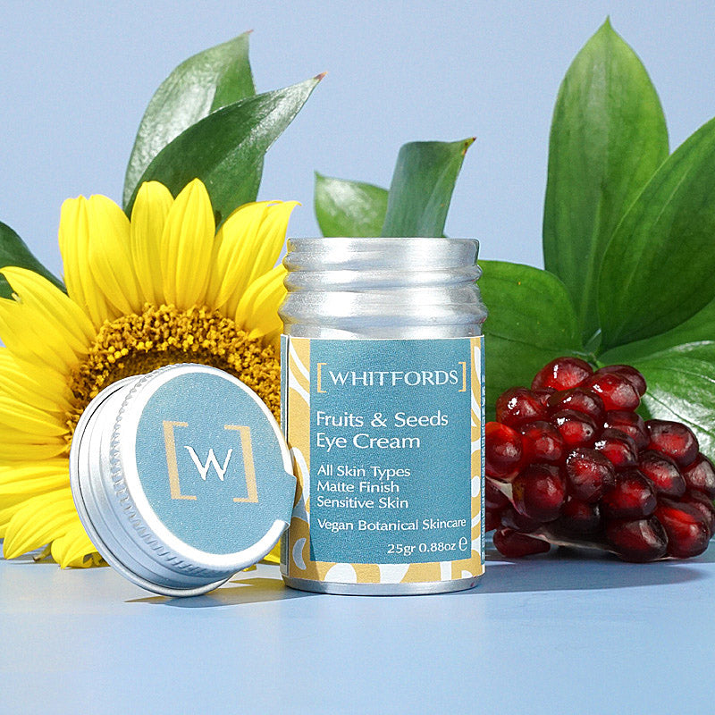 whitfords skincare fruits and seeds eye cream is pictured in a silver aluminium tin with a teal label which reads 'whitfords fruits and seeds eye cream. all skin types. matte finish. sensitive skin. vegan botanical skincare' the lid of the pot is leaning against the tin and in the background there is a sunflower, some green leaves and pomegranate seeds