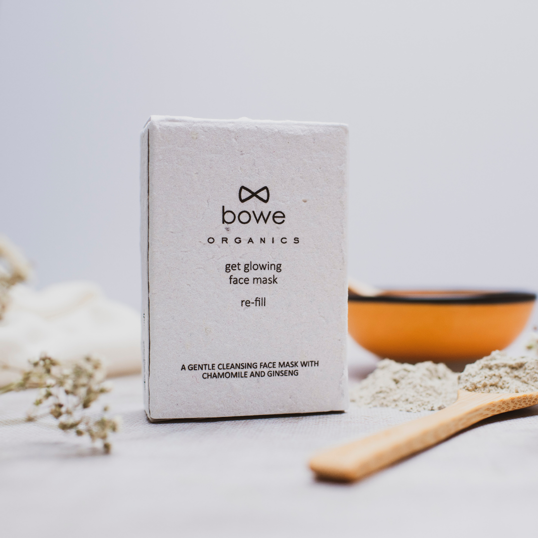 bowe organics face mask refill box can be seen in the foreground of a white/grey backdrop. There are some dried chamomile stems out of focus and an orange bowl with a wooden spoon in front of it which has some of the clay face mask powder on piled on it