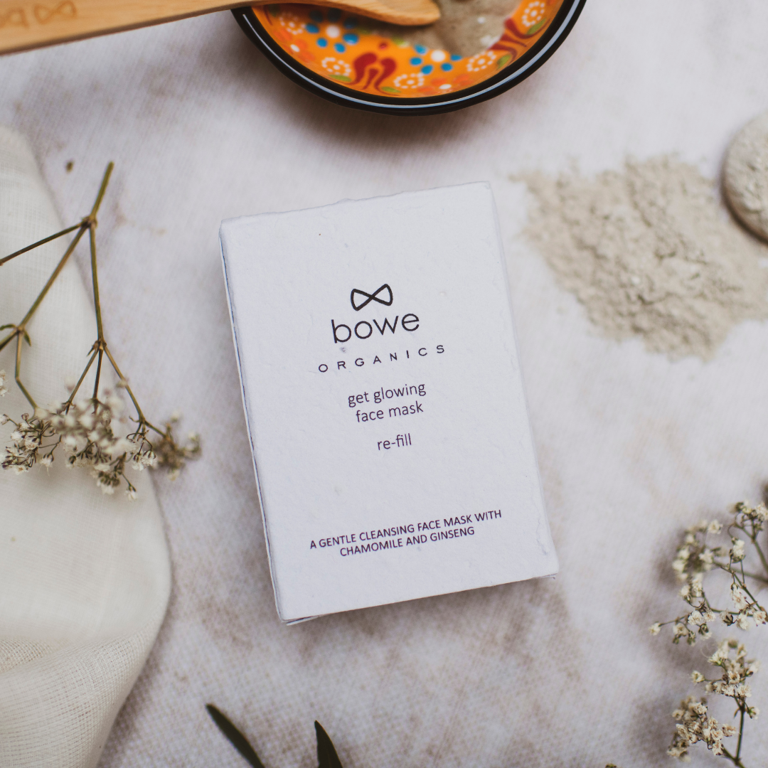 bowe organics glowing face mask refill box is photographed laid flat on a grey textured cloth. gypsophilia flowers and an orange hand painted bowl with a black rim and turkish read, green and blue pattern inside can be seen around the box and a pile of the powder mask