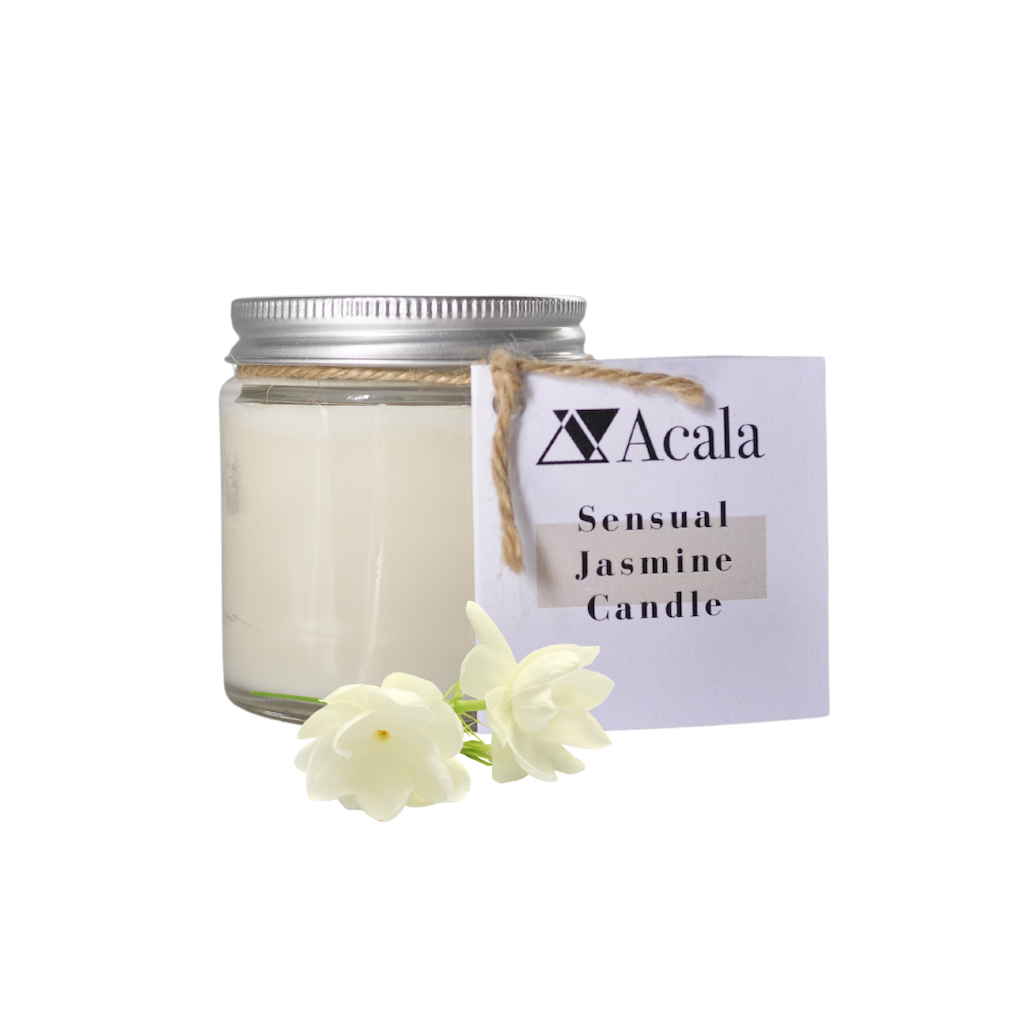 photo of acala zero waste's sensual jasmine candle on a white background. the candle comes in a clear glass jar with silver aluminium lid. there is some brown string which has a white card label attached to it which reads 'acala sensual jasmine candle'. in front of the candle are two buds of jasmine flowers