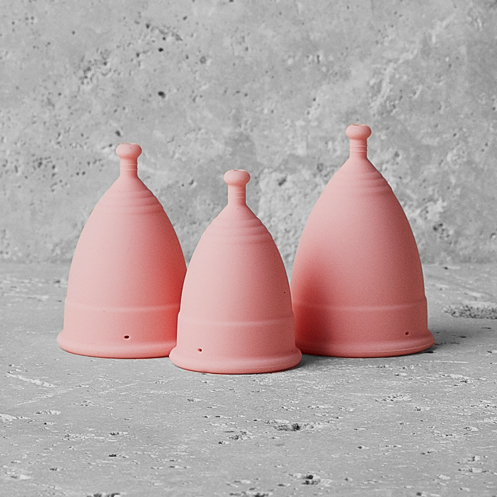& Sisters Nudie Period Cup. Sustainable menstrual care. All 3 sizes of the pink menstrual cup are shown on a concrete surface.