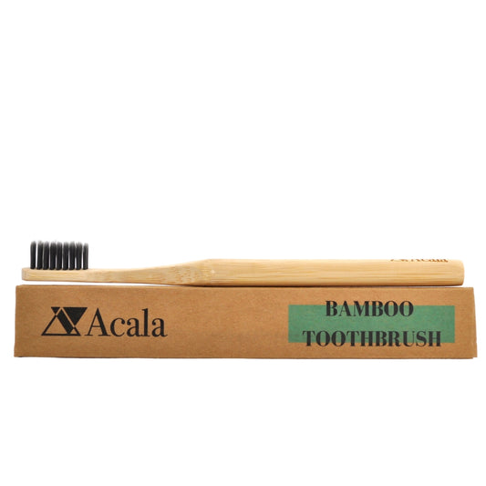 compostable and biodegradable bamboo toothbrush with dark grey charcoal bristles lying flat on top of a brown kraft paper toothbrush box which has the acala zero waste shop's logo printed on it in black text with a dark turquoise accent