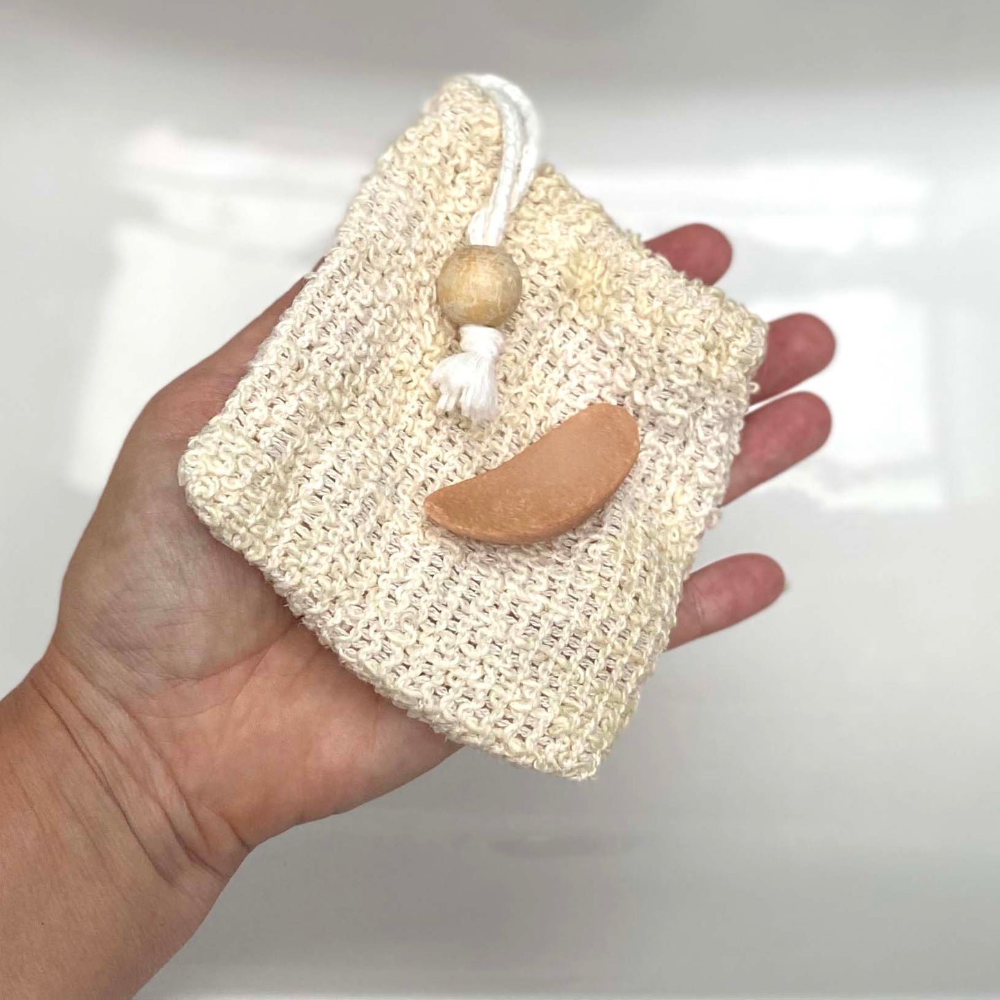 A small piece of pink soap placed on top of the natural fibre soap pouch being held by a hand against a white background