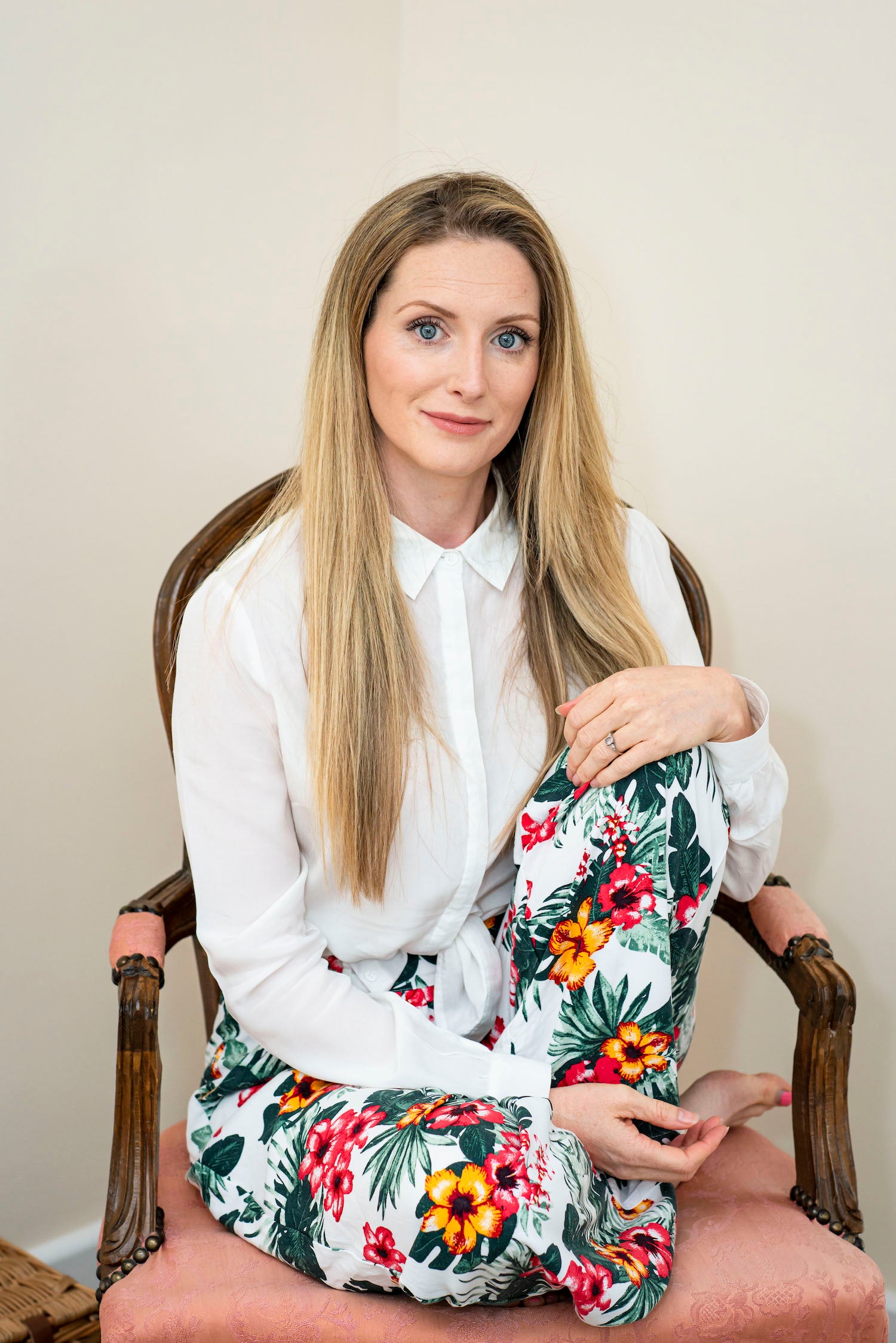 Bowe Organics Founder Diane Bowe. Diane is sitting on a wood chair with pink upholstery. Her long blonde hair is down over a white collared shirt and she is wearing colourful floral trousers.