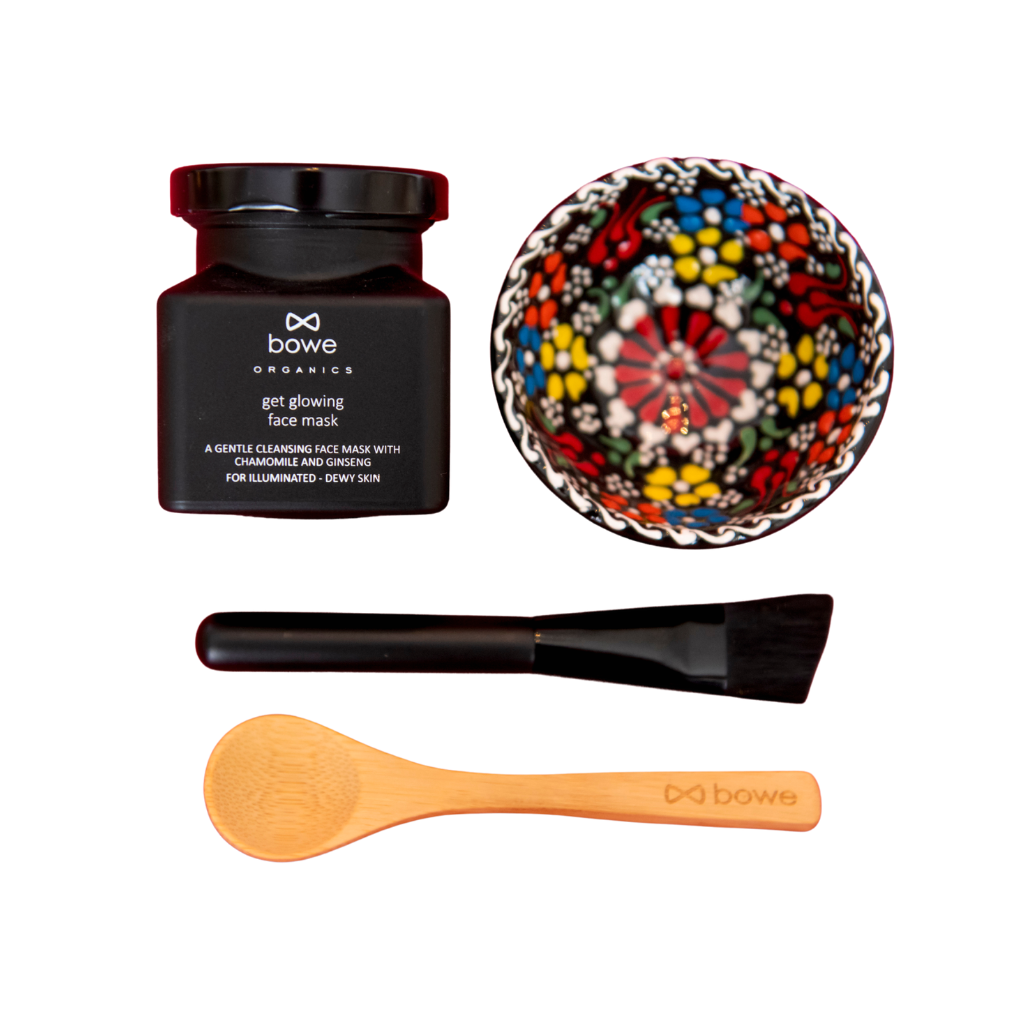 Flat lay of the products that are in the bowe organics get glowing face mask skincare gift set. Clockwise from left: a black frosted glass jar with a clay powder face mask inside for oily or combination skin. Black, blue, yellow, white and red hand painted floral bowl in turkish style. Black synthetic vegan and cruelty free face mask applicator brush. Wooden spoon with bowe organics brand name and infinity logo in the shape of a bow engraved on it