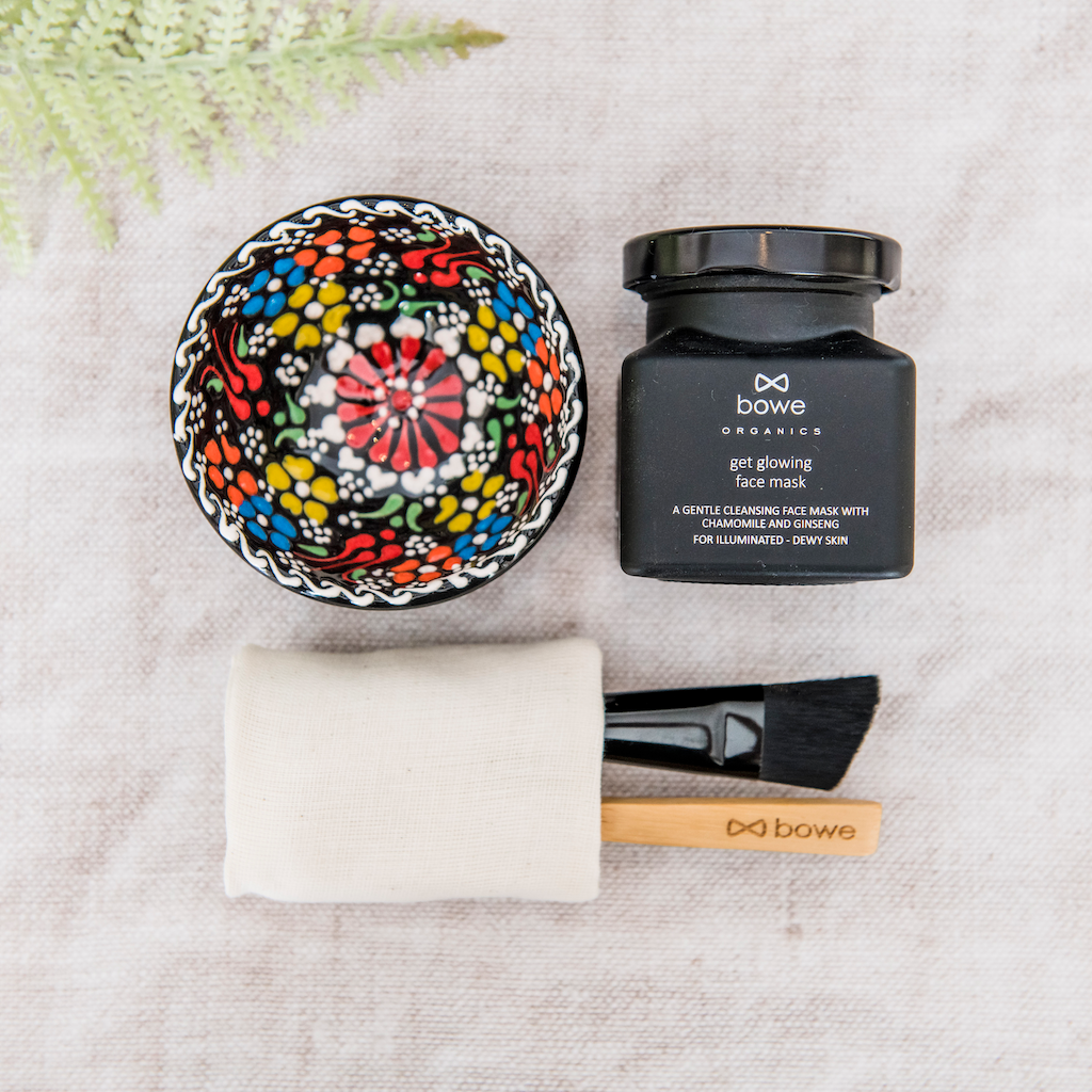 flat lay of natural skincare products on a linen cloth with a light green fern in the background. The face mask kit includes, clockwise from left a hand painted turkish bowl which is black, orange blue, green, white and yellow in this photo. A black glass jar with metal lid containing a green clay, chamomile and ginseng powder face mask. A white organic cotton cloth is wrapped around the face mask applicator brush and wooden mixing spoon