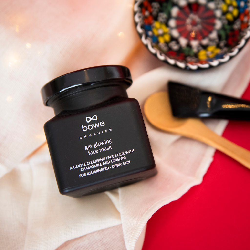 the bowe organics get glowing face mask black jar is displayed on top of the white gots organic cotton face flannels which are displayed open on a red surface. you can see the hand painted face mask bowl, brush and spoon in the background blurred