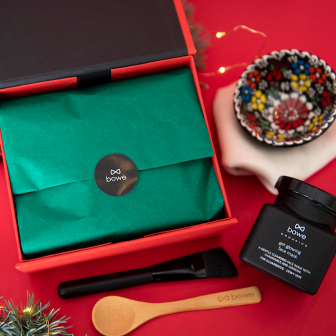 glowing clay chamomile and ginseng face mask photographed on a red background. You can see the red gift box with dark green tissue paper inside and laid around the box is the handpainted turkish style bowl in black sitting on top of an organic cotton muslin cloth. The black face mask jar is in front of the mask box and next to the face mask brush and wooden spoon. there are lights and fir branches in the background