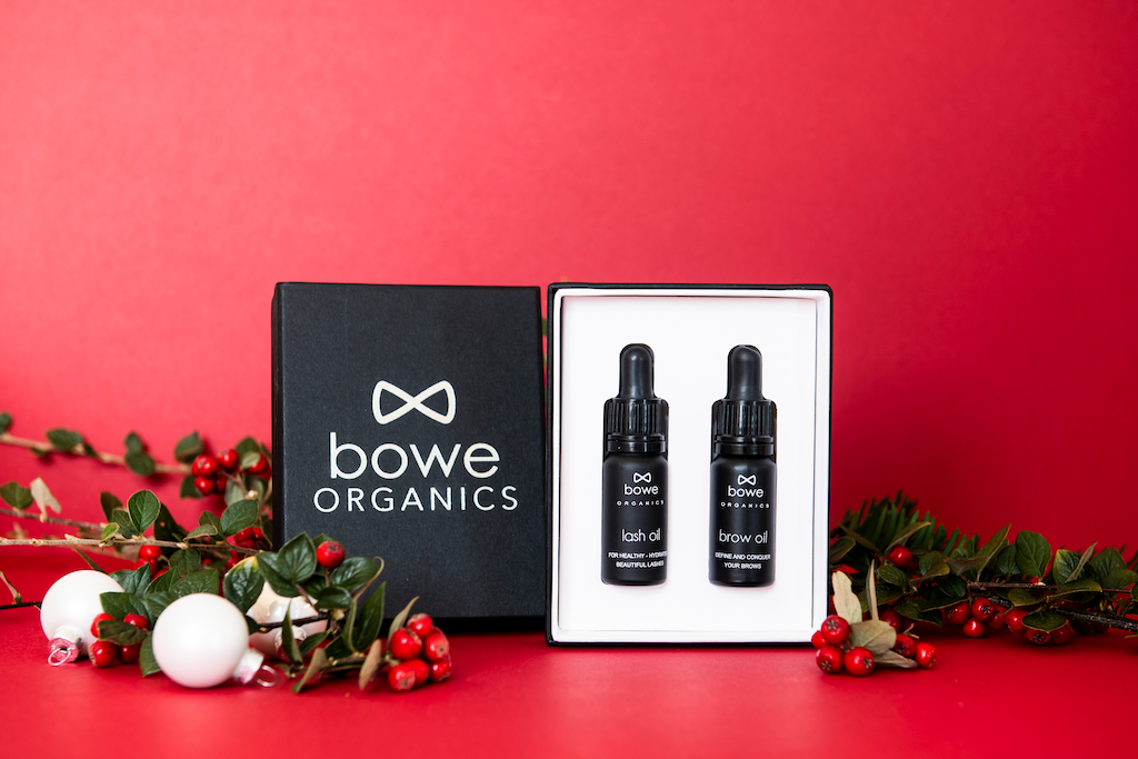 Bowe Organics Gift of Brows and Lashes. Vegan Christmas gifts. The lash and brow oil are pictured in their box next to the silver embossed lid, on a red background with holly draped around.