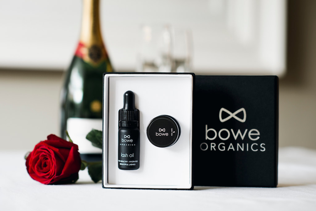 Bowe Organics Gift of Lips and Lashes. Vegan Valentine's gift. The lash oil and lip balm are pictured in their box with the silver embossed lid next to it, a single rose stem next to it, and a bottle of champagne out of focus in the background.