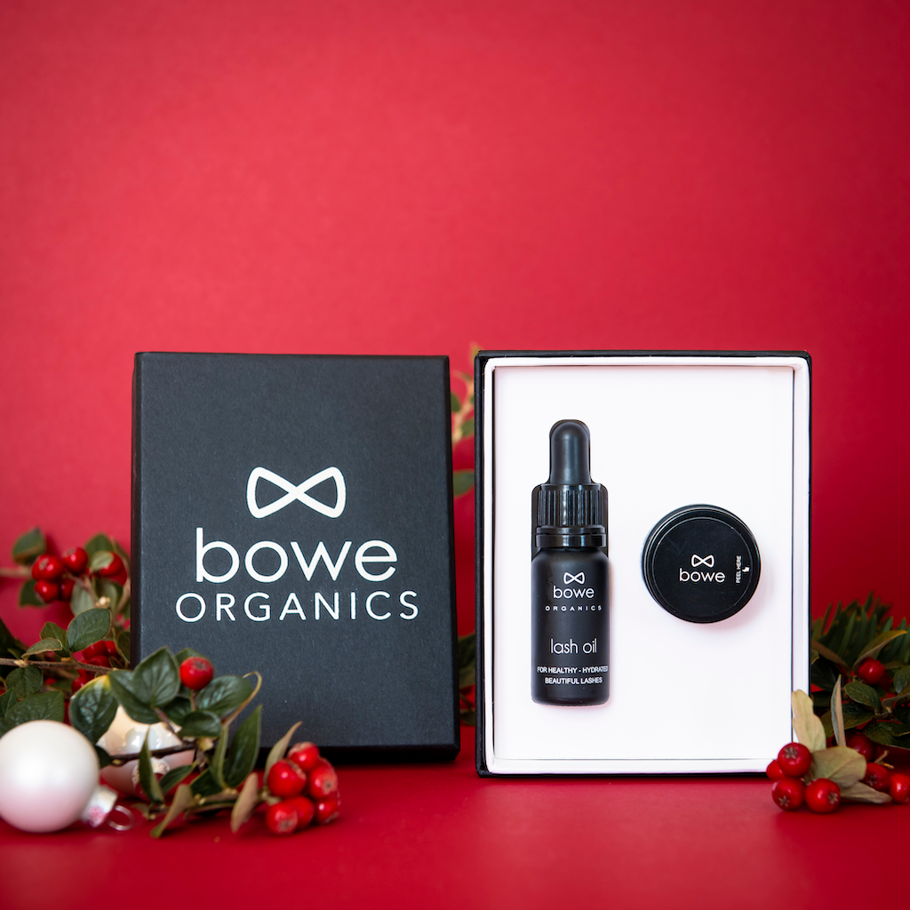 Bowe Organics Gift of Lips and Lashes. Natural Christmas gifts. The lash oil and lip balm are pictured in their box with the silver embossed lid next to it, and some holly branches draped around on a red background.