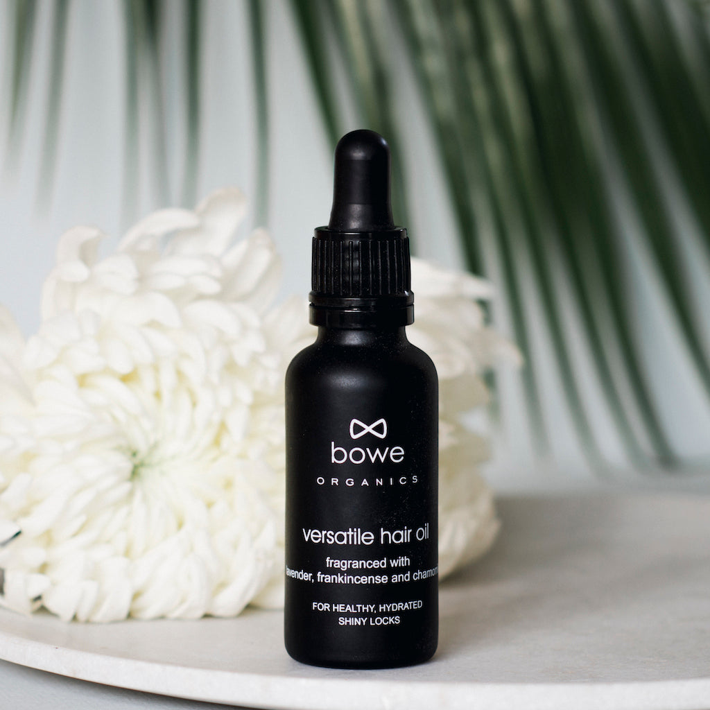 Bowe Organics Versatile Hair Oil Fragranced. Vegan hair treatment. The oil is pictured in a black glass bottle with white text and a black pipette. There is a white flower and a palm leaf in the background.