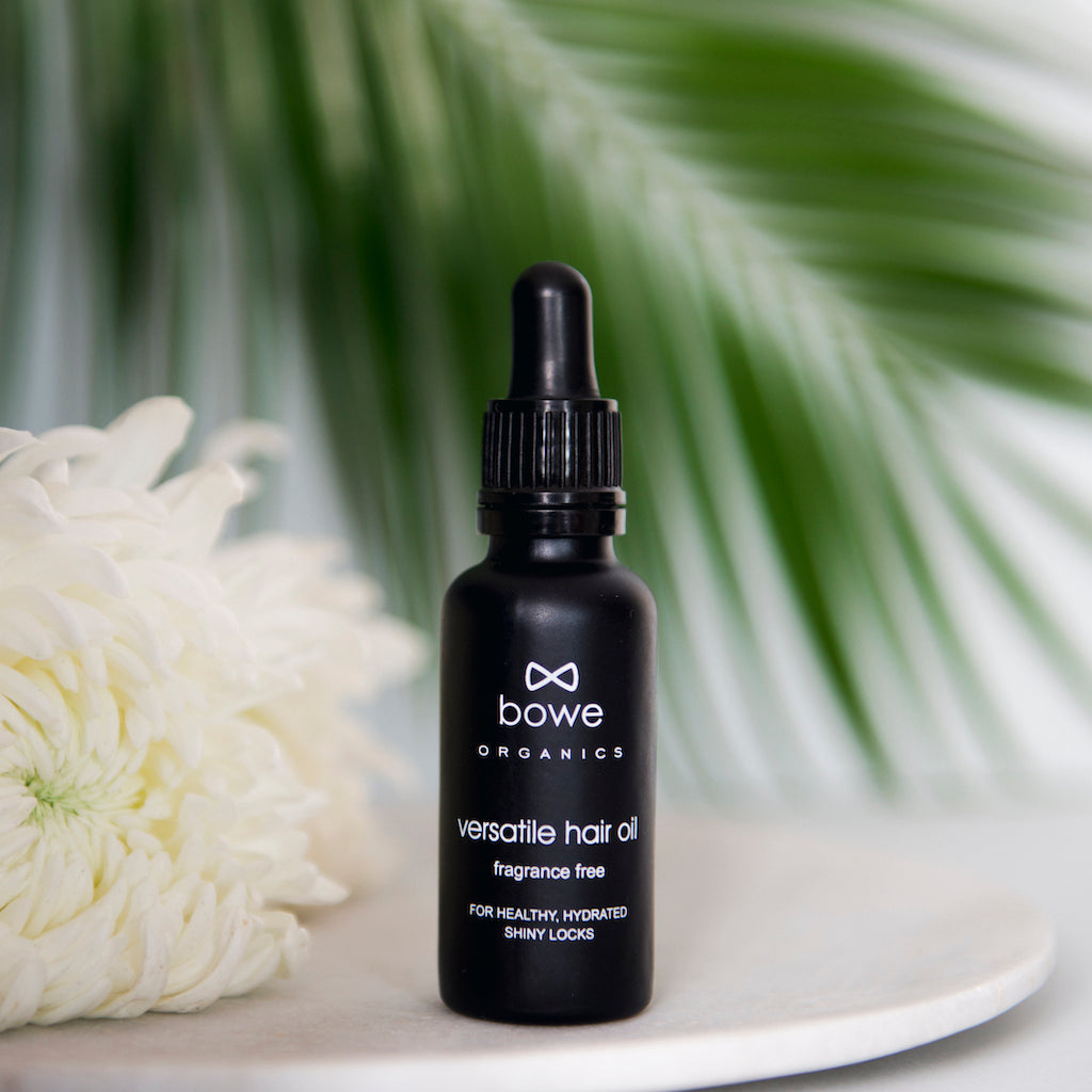 Bowe Organics Versatile Hair Oil Fragrance Free. Vegan pre shampoo treatment. The oil is pictured in a black glass bottle with white text and a black pipette. There is a white flower and a palm leaf in the background.
