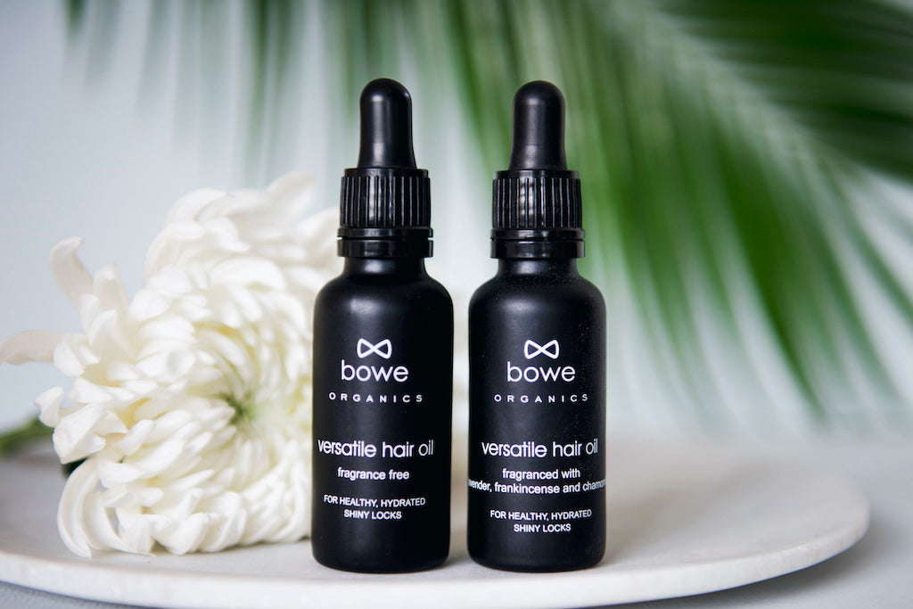 Bowe Organics Versatile Hair Oils. Natural pre shampoo treatment. The oils are pictured in black glass bottles with white text and black pipettes. There is a white flower and a palm leaf in the background.