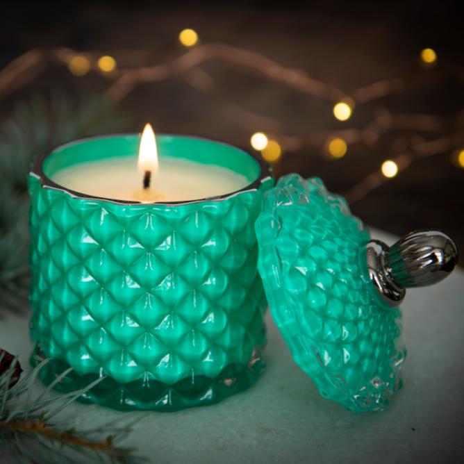 green candle jar with lit candle and background fairy lights next to fir branch
