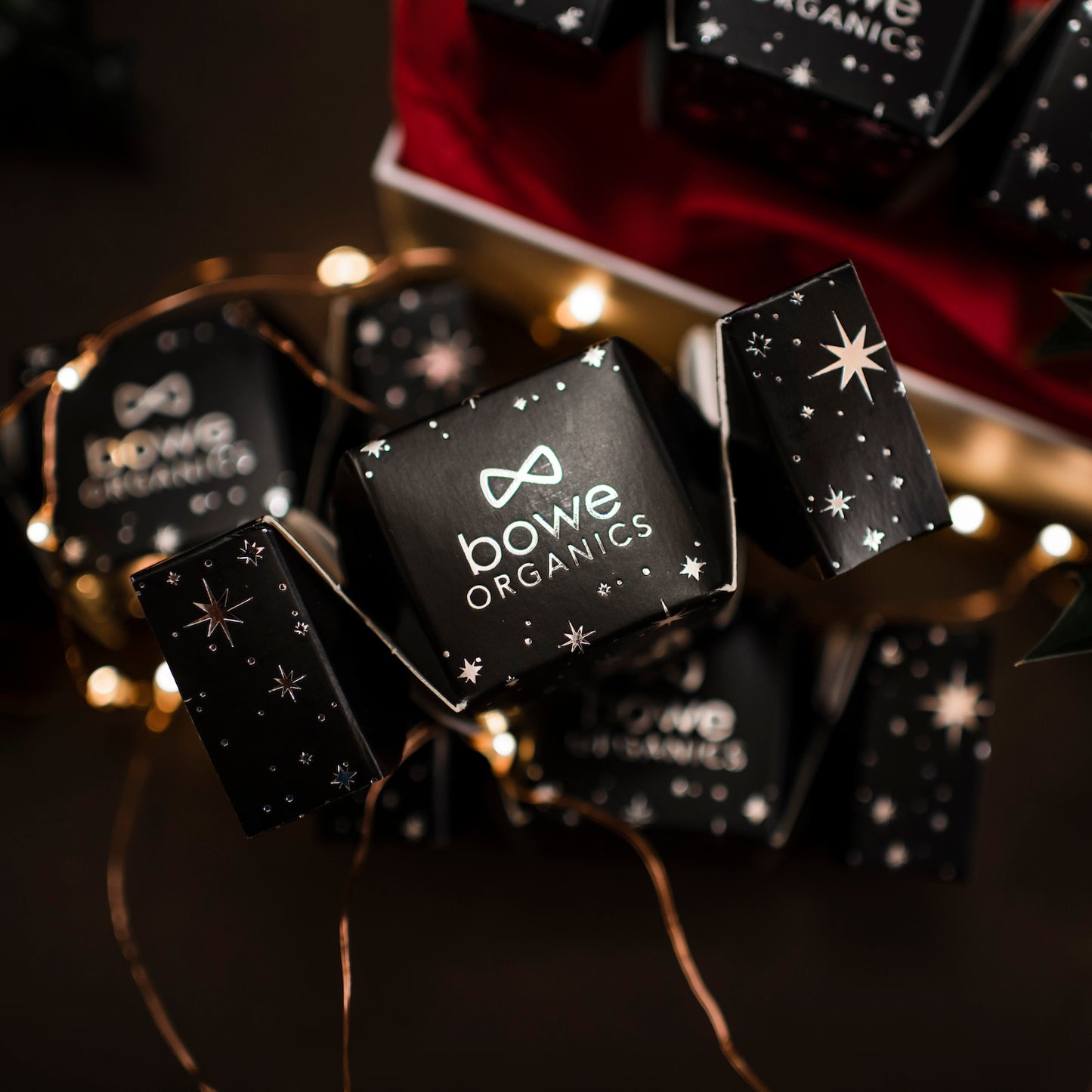 A small black Bowe Organics christmas cracker with white stars and sparkles set against a dimly lit background with more crackers and fairy lights in the background.