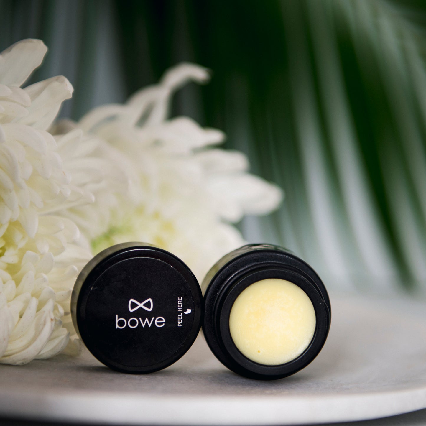 Bowe Organics lip rescue balm photographed with blurred out green leaves and white chrysanthemums in the background.  The pot is open facing towards camera with the lid next to it and showing the bowe organics name and logo