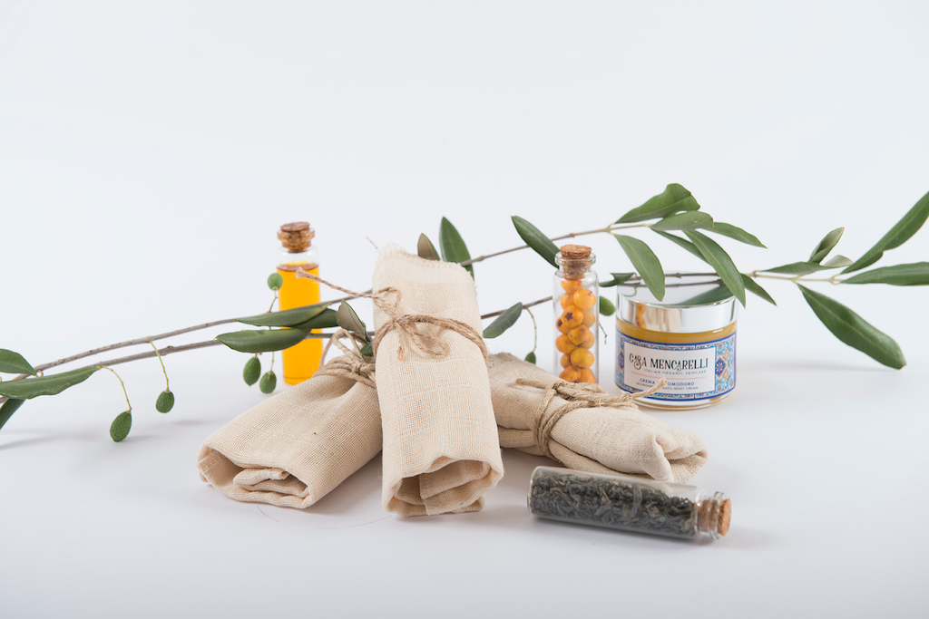 Casa Mencarelli Organic Cotton Face Cloths. Certified organic cotton face cloths. The three cotton cloths are pictured rolled up with twine and stacked in a fan shape. There are branches with small leaves draped behind and a pot of one of Casa Mencarelli's balms next to them.