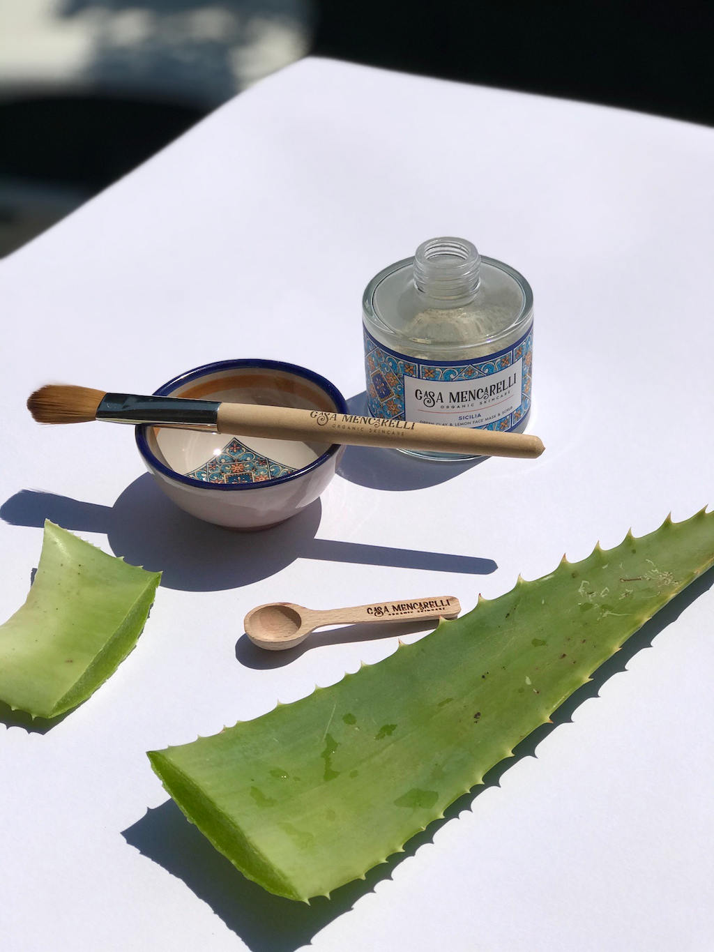 Casa Mencarelli Sicilia Green Clay & Lemon Face Mask & Scrub. Vegan face scrub. The mask jar is pictured next to an empty Casa Mencarelli mask bowl with a mask brush sitting on top and a cut aloe leaf next to them.