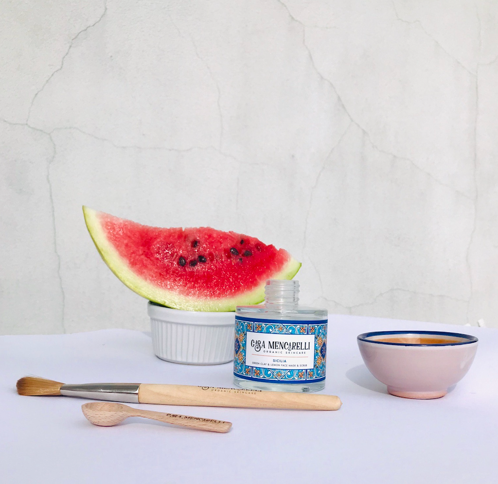 Casa Mencarelli Sicilia Green Clay & Lemon Face Mask & Scrub. Certified organic scrub. The glass jar is pictured open next to a mask bowl and brush, along with a slice of watermelon behind.