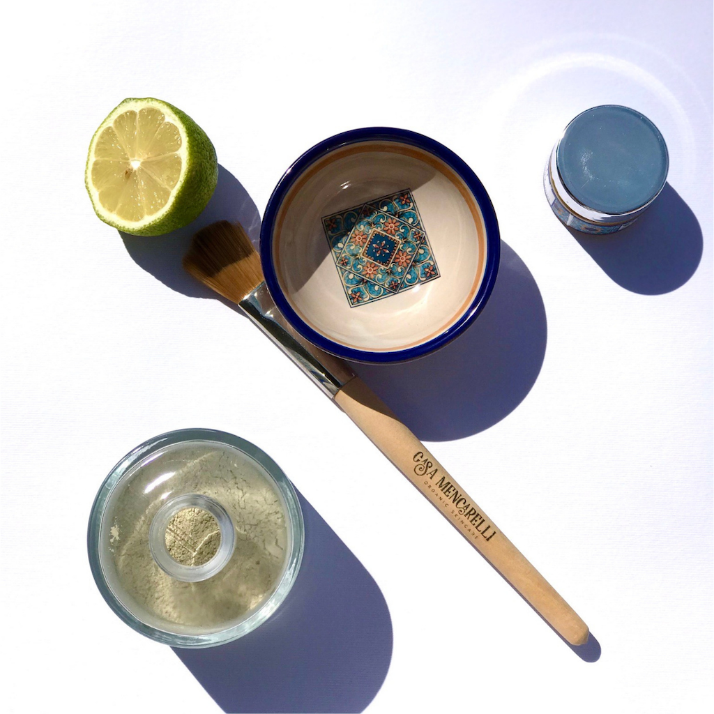Casa Mencarelli Face Mask Brush. Sustainably sourced face mask brush. The wooden mask brush is branded on the stem with the Casa Mencarelli logo. It is sitting next to a Casa Mencarelli mask bowl, a sliced lime, and an open jar of Casa Mencarelli face mask all pictured from above.