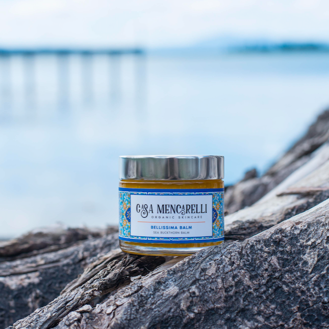 Casa Mencarelli Bellissima Sea Buckthorn Balm. Multipurpose natural skincare. The balm is pictured in a glass jar and labelled with illustrations of the traditional tiling of Umbria in Italy. It is sitting on a stretch of tree root with a body of water visible in the background.
