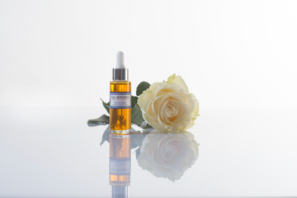 Casa Mencarelli Sole E Mare Sea Buckthorn Serum. Vegan face massage oil. The orange serum is packaged in a clear glass bottle with a label decorated with illustrations of traditional tiling from Umbria in Italy. A white rose is sitting next to the bottle and they are sitting on a reflective surface.