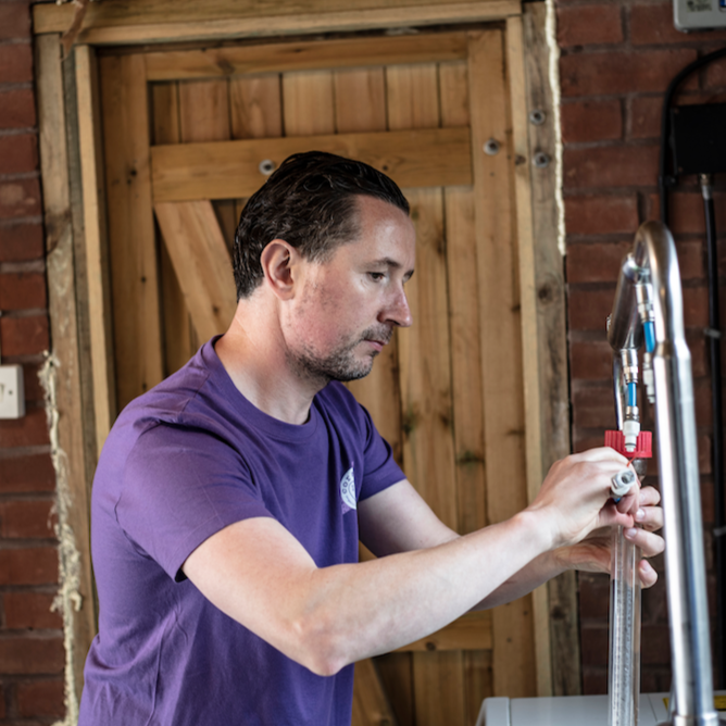 kevin, coraline skincare's founder working in his soapery in devon. he is distilling some essential oils and behind him is a red brick wall with wooden panelled door