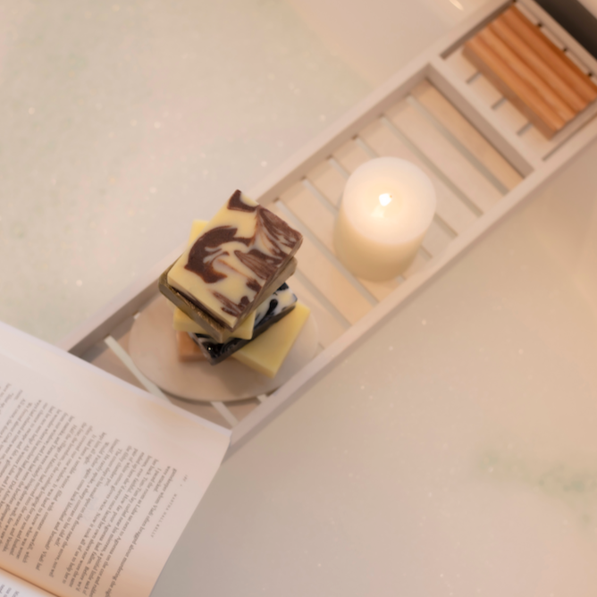 aerial view of a white bath with bubbles. Across the bath is a white wooden tray which has a stack of multicoloured hand made and handcut soaps on it. a white candle which is lit and a book. Inspiration for a relaxing bath routine with coraline's cinnamon swirl soap