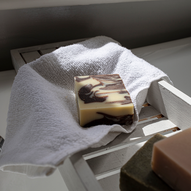 bar of cinnamon soap which is sat on top of a fluffy white face cloth in a wooden tray that sits across a white bath tub. 