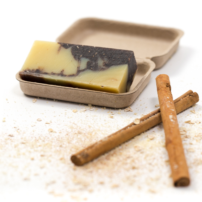 slice of creamy chocolate coloured soap in a brown cardboard tray with ground almonds and cinnamon sticks displayed next to it