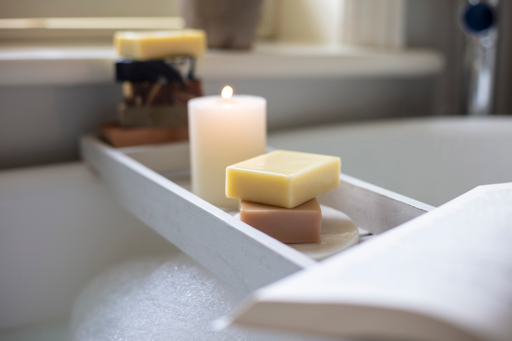 A serene bath setup featuring a wooden tray over a bubble bath with assorted handmade soap bars and a lit candle, creating a relaxing and cozy spa-like atmosphere.