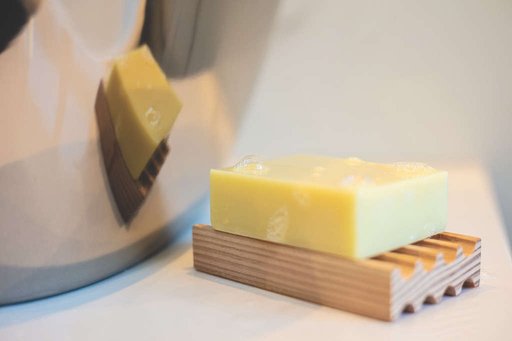 A bar of yellow soap on a small wooden soap dish, with a blurred reflection of the soap visible on the shiny surface of a kettle. the background is soft and pale.