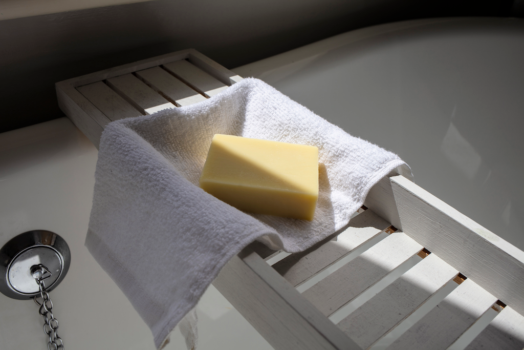 A yellow bar of soap rests on a white towel, which is placed on a wooden bath caddy over a bathtub, illuminated by natural light.