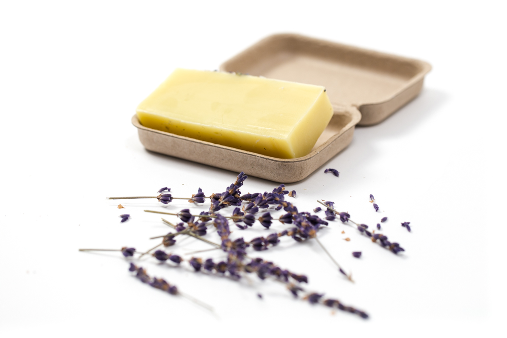 A bar of yellow soap on a beige tray next to dried lavender sprigs, all set against a white background.