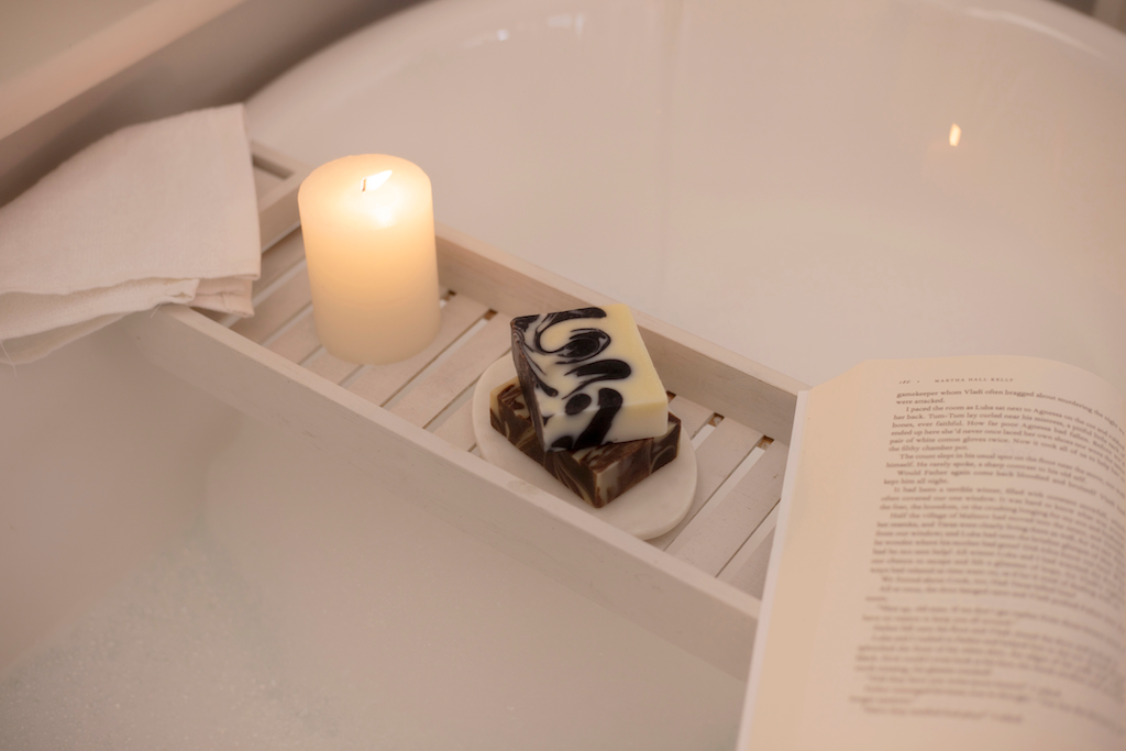 A relaxing bathtub scene features a lit white candle, a bar of the minty fresh soap, and an open book positioned on a wooden bath tray. A folded white towel hangs from the tray's side, and the tub is filled with soapy water.