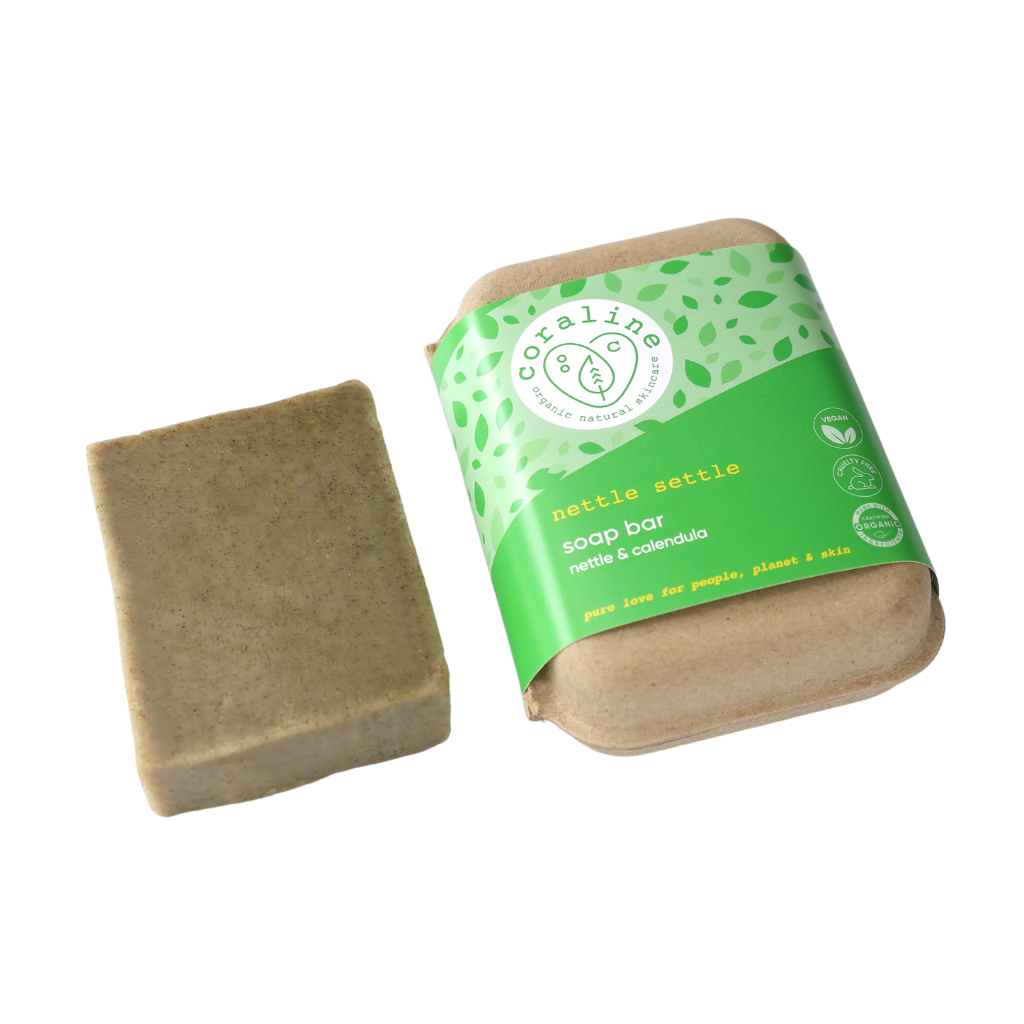 The Coraline nettle settle green soap bar lies next to its eco-friendly paper packaging. The packaging is partially green with leaf patterns and reads "Coraline, nettle settle soap bar, nettle & calendula - pure love for people, planet & skin.