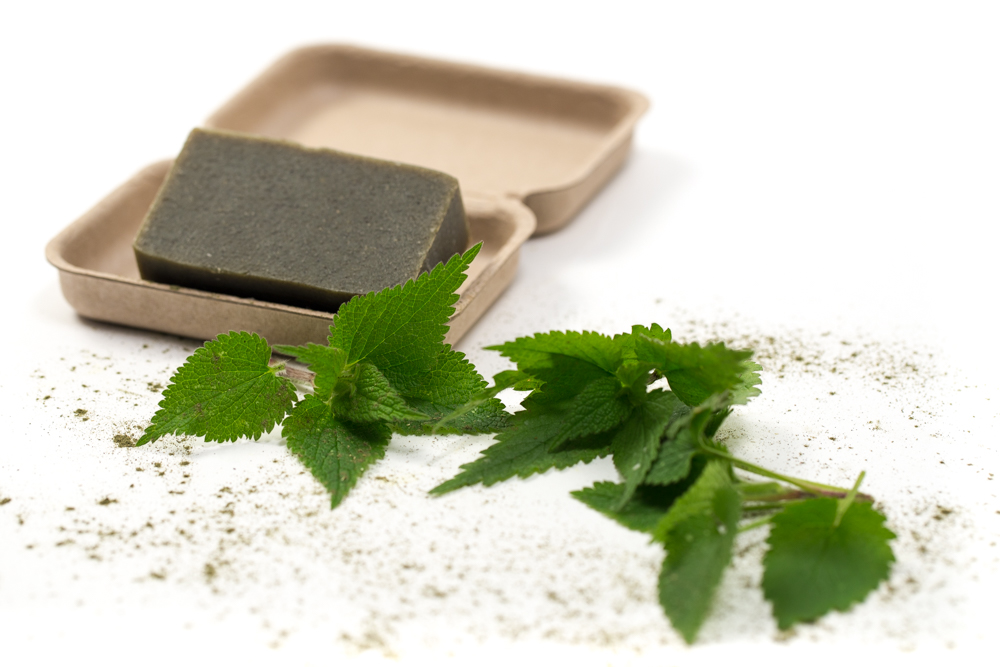 A green/brown soap bar rests on a wooden soap dish against a white background. In the foreground, fresh green nettle leaves are scattered alongside some nettle powder.