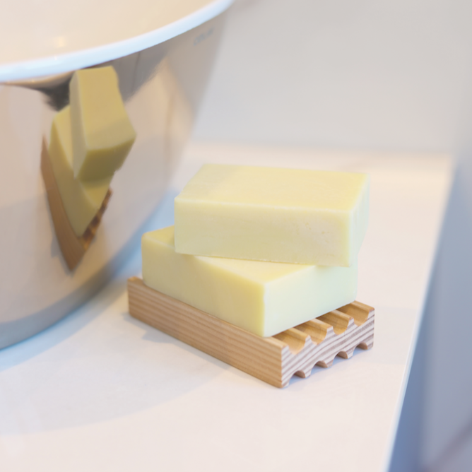 wooden soap rack sits on top of a white bathroom counter and has creamy white soaps stacked on top of it. you can see the reflection of the soaps and the dish in the chrome coloured basin to the left