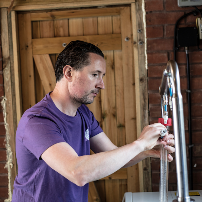 kevin, coraline skincare's founder working in his soapery in devon. he is distilling some essential oils and behind him is a red brick wall with wooden panelled door