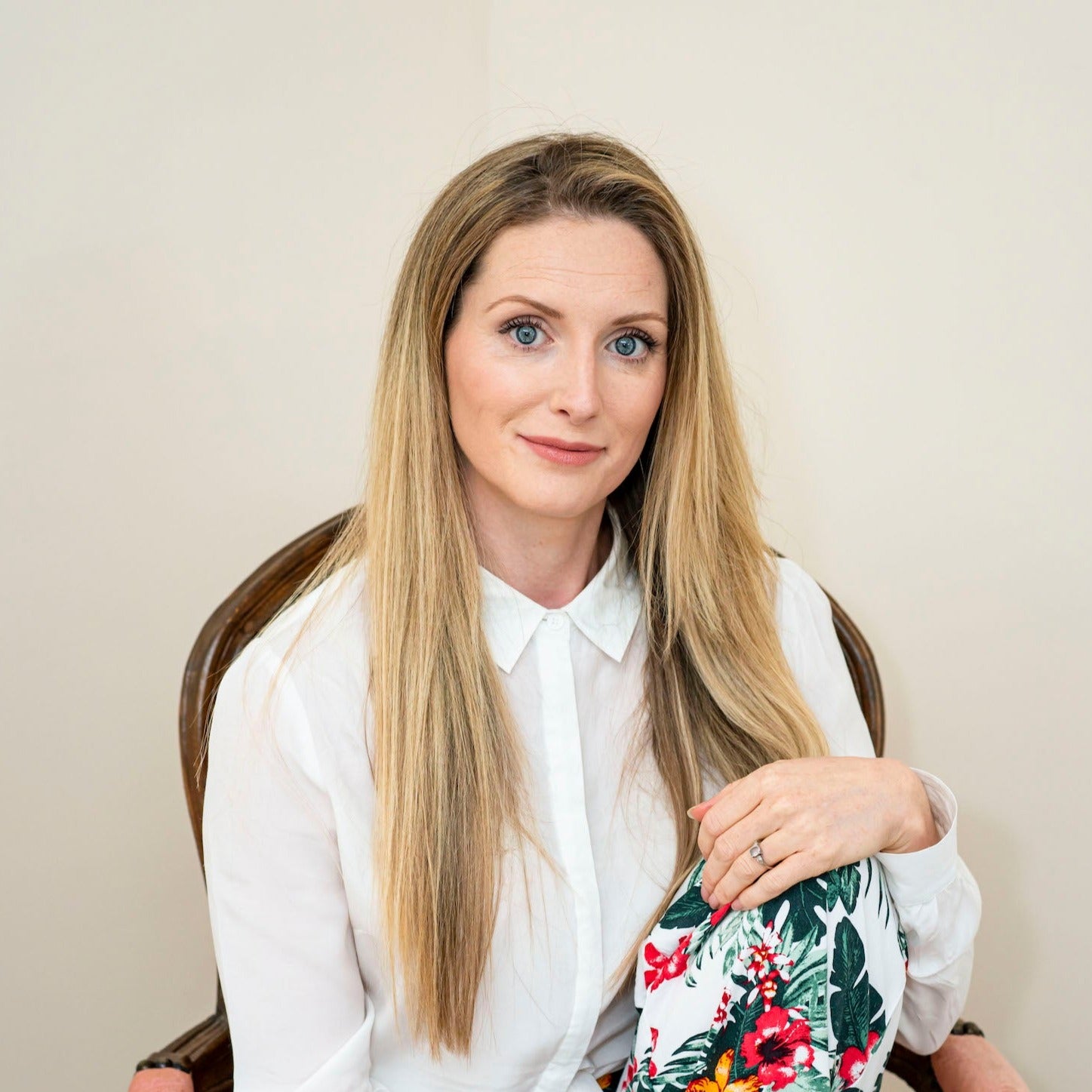 Bowe Organics Founder Diane is photographed sitting in a wooden chair with arm rests and a coral fabric. Diane has long blonde hair and bright blue eyes and she's seen with one leg raised up to her torso in a sitting position with her hand resting on her knee. Diane is wearing a white collared shirt and printed trousers with tropical flowers on them