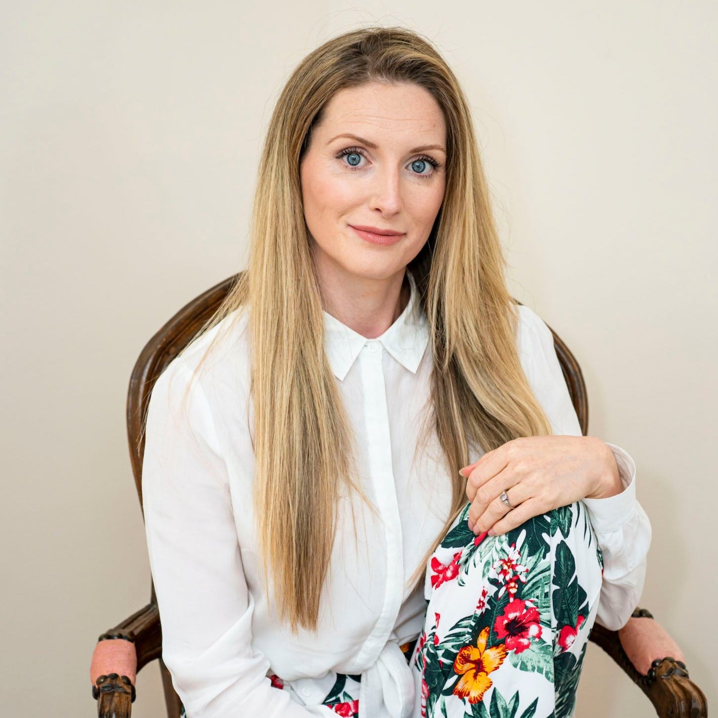 Bowe Organics Founder Diane is photographed sitting in a wooden chair with arm rests and a coral  fabric. Diane has long blonde hair and bright blue eyes and she's seen with one leg raised up to her torso in a sitting position with her hand resting on her knee. Diane is wearing a white collared shirt and printed trousers with tropical flowers on them