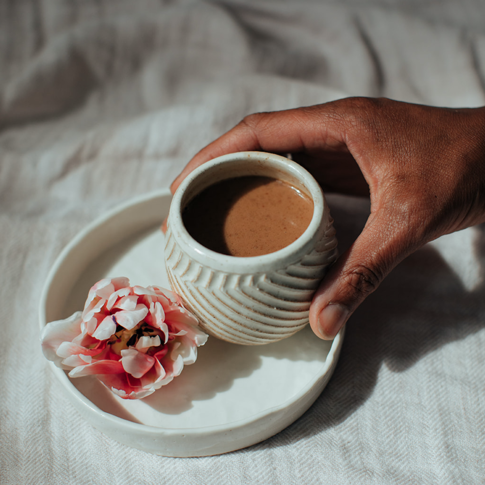 Forage Botanicals Aunt Flo's Raw Drinking Chocolate. Vegan iron supplement. The cacao drink is pictured in a ceramic mug without a handle, held in a person's hand, on top of a larger white ceramic tray with a pink flower on it.