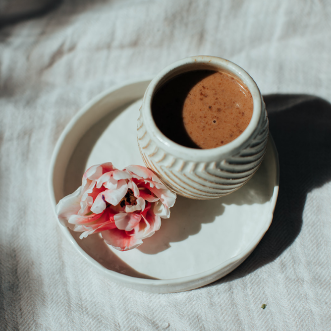 Forage Botanicals Aunt Flo's Raw Drinking Chocolate. Vegan iron supplement for menstruation. The cacao drink is pictured in a ceramic mug without a handle, on top of a larger white ceramic tray with a pink flower on it.