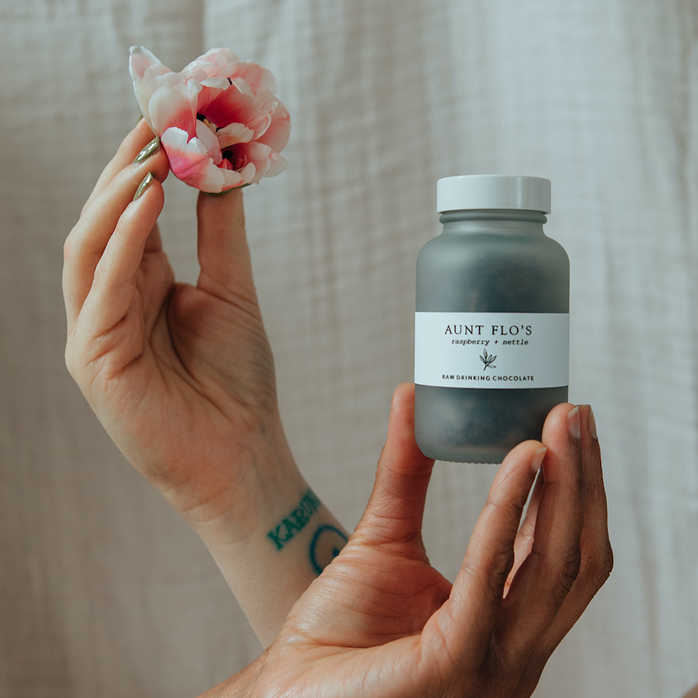 Forage Botanicals Aunt Flo's Raw Drinking Chocolate. Natural iron supplement for hormonal cycles. The glass jar filled with raw cacao is being held by one person's hand, while another person's hand holds a pink flower behind.