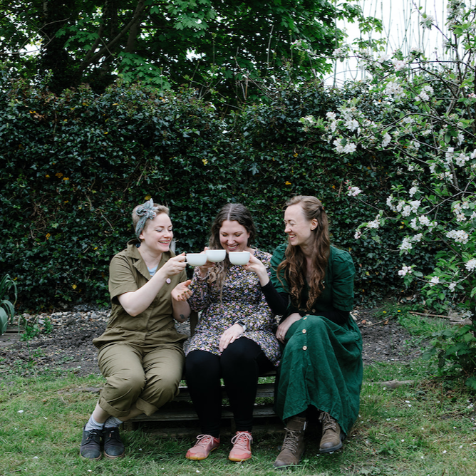 Forage Botanicals Aunt Flo's Raw Drinking Chocolate. Vegan iron supplement for anemia. Forage founder Natasha is sitting on the left with two other people enjoying a cuppa. They are sitting in a flourishing garden on a bench.