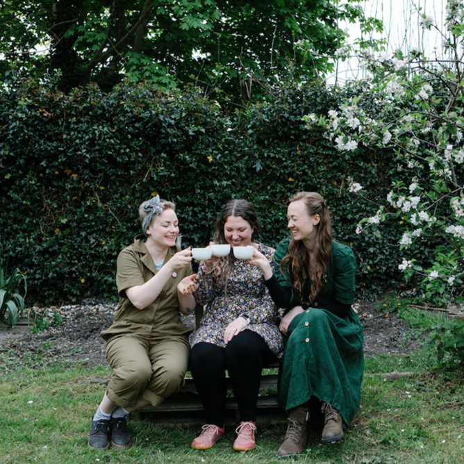 natasha richardson author, medical herbalist and founder of forage botanicals is sitting on the left of 3 women on a wooden bench in a garden with green shrubbery. The women are all dressed in green and brown tones and are gently pressing their white tea cups together in a cheers