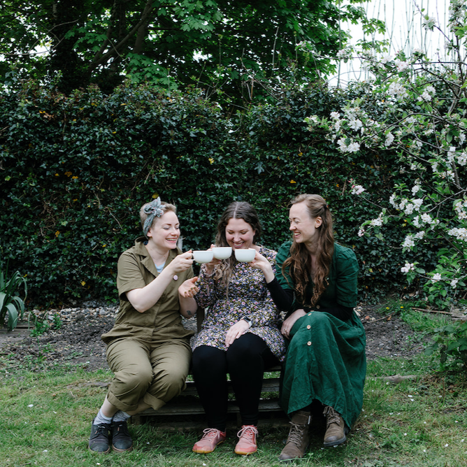 natasha richardson author, medical herbalist and founder of forage botanicals is sitting on the left of 3 women on a wooden bench in a garden with green shrubbery. The women are all dressed in green and brown tones and are gently pressing their white tea cups together in a cheers
