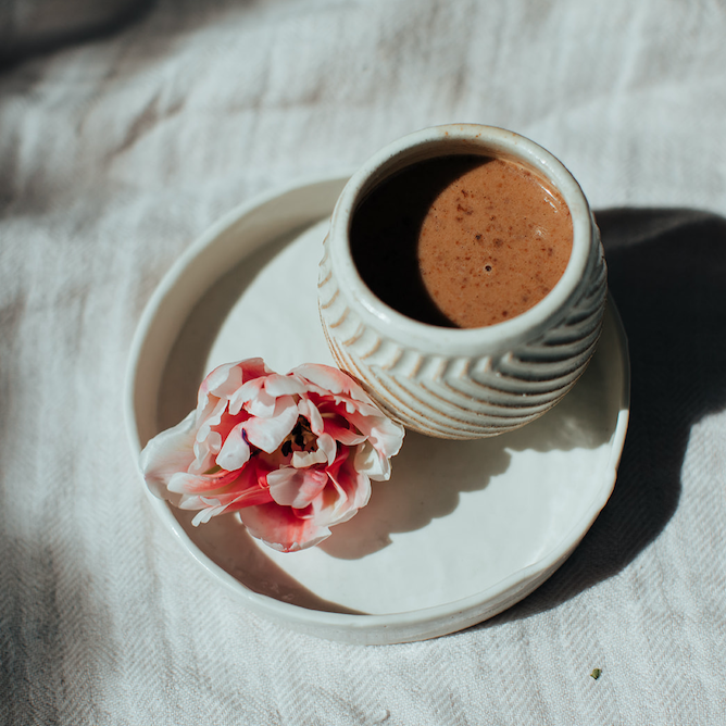 rich chocolatey coloured raw cacao drink is in a white cup with no handle and some arrow textures and patterns on it. Sitting on a white saucer with a pink and white peony flower 