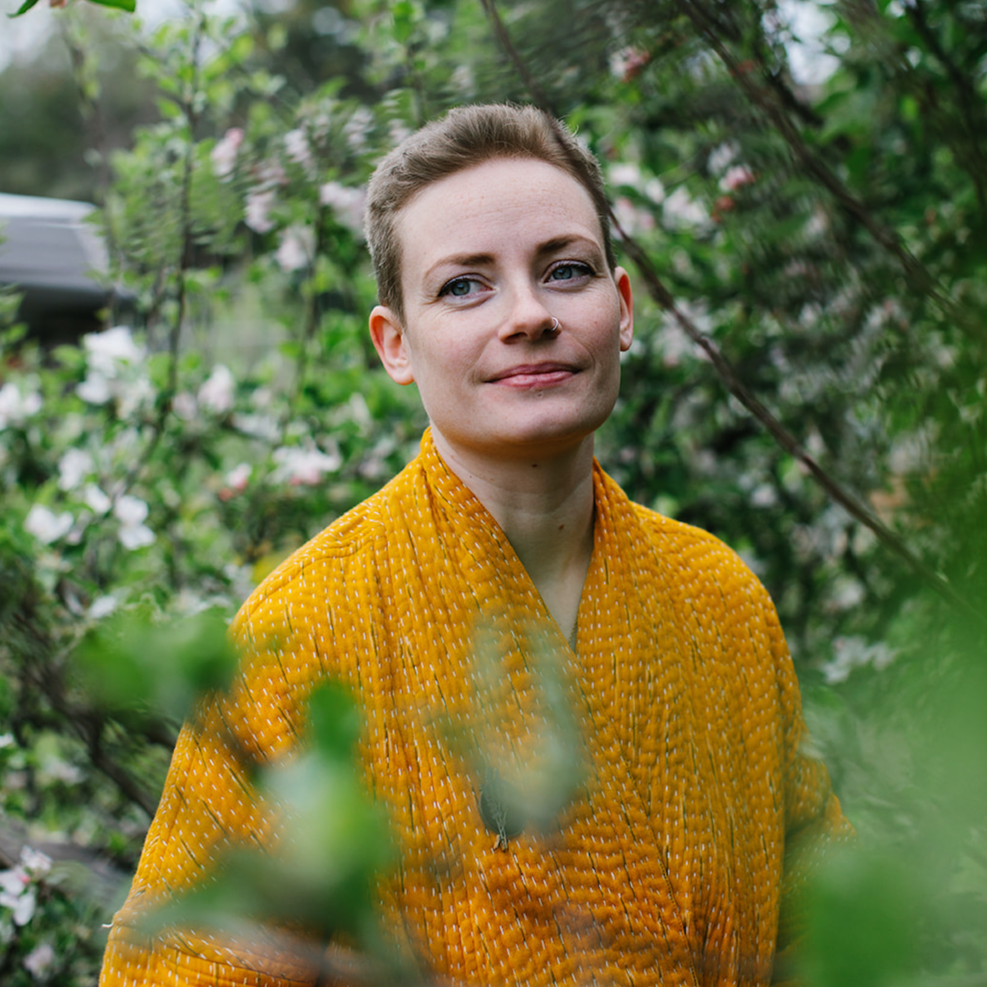 natasha richardson is a medical herbalist and founder of hormonal support products range forage botanicals. she is photographed amongst green shrubbery with white blossoms on it wearing an orange yellow jacket. she has short strawberry blonde hair, blue eyes and pale skin